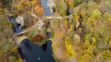 Beautiful scenery view of autumn park with trees with yellow fallen leaves, lakes, architecture, glades and people walking along dirt paths on autumn day.Top view. Flying over the autumn park.