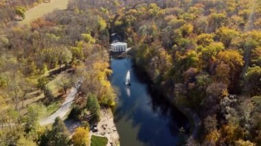 Beautiful panoramic autumn landscape park, many trees with yellow leaves, lake with fountain in center, architecture, big stones, paths walkways, people walking on sunny autumn day. Aerial drone view