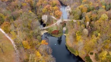 Flying over lakes, trees with yellow red green orange fallen leaves and people walking on dirty paths. Top view. Landscape aerial view autumnal park. Large beautiful landscaped park in autumn day.