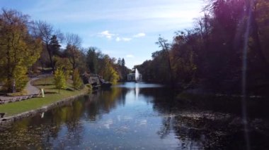 Beautiful landscaped park with decorative fountain in center of lake, trees and large stones on banks on sunny autumn day. Flying over water with fallen leaves and blue sky reflection. Many tourists