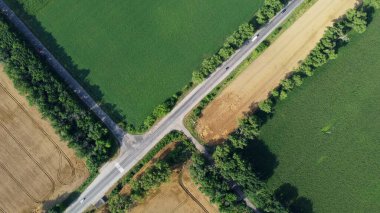 Automobile asphalt road with cars driving along it between agricultural fields with yellow ripened wheat and different green agricultural crops. Aerial drone view. Agricultural agrarian crop landscape