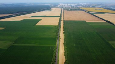 Farklı tarımsal yeşil bitkiler, olgun sarı buğdaylar, hareket halindeki arabaların ve şehrin bulunduğu güzel panoramik manzara. - Hava aracı. Tarım mahsulü manzarası