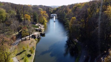 Göllü, çeşmeli, ağaçlı, büyük taşlı, güneşli bir sonbahar gününde yürüyen ve beyaz mimarisi olan güzel bir park. Sahilde sarı yapraklı ağaçlar, yüksek akarsu akıntısı olan turistler...