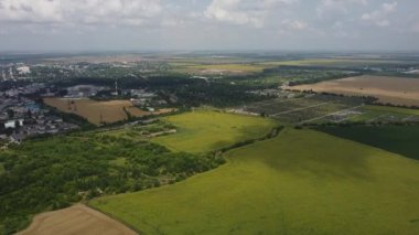 Tarım arazisi, çiçek açan büyük ayçiçekleri, yeşil tarım sanayi tarlaları, olgun buğday tarlaları, ağaçlar, bulut gölgeleri ve yaz güneşli günlerinde şehir binaları. Hava aracı perspektifi. Üst görünüm