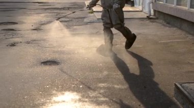 Koruyucu giysili bir adam şehrin kaldırımına antiseptik solüsyon sıkıyor. Koronavirüsü önlemek için sprey kimyasalların dezenfekte edilmesi. Halka açık yerlerde hijyenik önlemler. Koronavirüs salgını