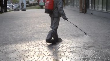 Koruyucu giysili bir adam şehrin kaldırımına antiseptik solüsyon sıkıyor. Koronavirüsü önlemek için sprey kimyasalların dezenfekte edilmesi. Halka açık yerlerde hijyenik önlemler. Koronavirüs salgını