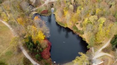 Gölü olan sonbahar parkı manzarası, gölde yüzen beyaz kuşlar, toprak yolda bisiklet süren iki kişi, kara yollarında yürüyen insanlar, sarı ve kırmızı yapraklı ağaçlar. Havadan