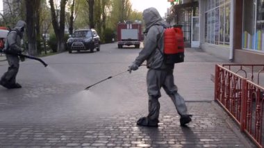 Koruyucu giysili bir adam şehrin kaldırımına antiseptik solüsyon sıkıyor. Koronavirüsü önlemek için sprey kimyasalların dezenfekte edilmesi. Halka açık yerlerde hijyenik önlemler. Koronavirüs salgını