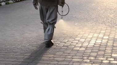 Koruyucu giysili bir adam şehrin kaldırımına antiseptik solüsyon sıkıyor. Koronavirüsü önlemek için sprey kimyasalların dezenfekte edilmesi. Halka açık yerlerde hijyenik önlemler. Koronavirüs salgını
