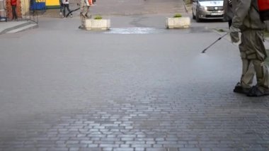 Koruyucu giysili bir adam şehrin kaldırımına antiseptik solüsyon sıkıyor. Koronavirüsü önlemek için sprey kimyasalların dezenfekte edilmesi. Halka açık yerlerde hijyenik önlemler. Koronavirüs salgını