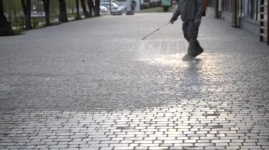 Koruyucu giysili bir adam şehrin kaldırımına antiseptik solüsyon sıkıyor. Koronavirüsü önlemek için sprey kimyasalların dezenfekte edilmesi. Halka açık yerlerde hijyenik önlemler. Koronavirüs salgını