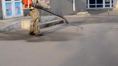 Koruyucu giysili bir adam şehrin kaldırımına antiseptik solüsyon sıkıyor. Koronavirüsü önlemek için sprey kimyasalların dezenfekte edilmesi. Halka açık yerlerde hijyenik önlemler. Koronavirüs salgını