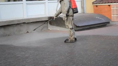 Koruyucu giysili bir adam şehrin kaldırımına antiseptik solüsyon sıkıyor. Koronavirüsü önlemek için sprey kimyasalların dezenfekte edilmesi. Halka açık yerlerde hijyenik önlemler. Koronavirüs salgını