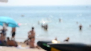 Güneşli yaz gününde arka planda bulanık deniz kumsalı ve birçok insan. İnsanlar kıyıda dinleniyor ve denizde yüzüyor. Güneş parıldıyor Bokeh. Tatil tatili, tatil gezisi, dinlenme ve dinlenme.