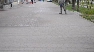 Koruyucu giysili bir adam şehrin kaldırımına antiseptik solüsyon sıkıyor. Koronavirüsü önlemek için sprey kimyasalların dezenfekte edilmesi. Halka açık yerlerde hijyenik önlemler. Koronavirüs salgını