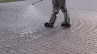 Koruyucu giysili bir adam şehrin kaldırımına antiseptik solüsyon sıkıyor. Koronavirüsü önlemek için sprey kimyasalların dezenfekte edilmesi. Halka açık yerlerde hijyenik önlemler. Koronavirüs salgını