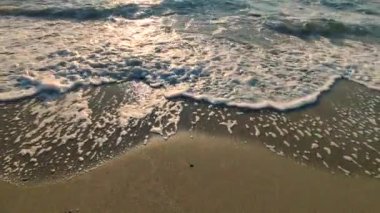 Gün batımında deniz kıyısındaki kumlu sahilde dalgalar dalgalanır. Beyaz köpüklü mavi deniz dalgaları kumsala geliyor. Deniz kıyısı kıyı şeridi. Doğal zemin, doğa zemin