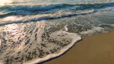 Gün batımında deniz kıyısındaki kumlu sahilde dalgalar dalgalanır. Beyaz köpüklü mavi deniz dalgaları kumsala geliyor. Deniz kıyısı kıyı şeridi. Doğal zemin, doğa zemin