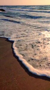 Gün batımında deniz kıyısındaki kumlu sahilde dalgalar dalgalanır. Beyaz köpüklü mavi deniz dalgaları kumsala geliyor. Deniz kıyısı kıyı şeridi. Doğal arka plan, doğa. Dikey