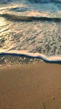 Gün batımında deniz kıyısındaki kumlu sahilde dalgalar dalgalanır. Beyaz köpüklü mavi deniz dalgaları kumsala geliyor. Deniz kıyısı kıyı şeridi. Doğal arka plan, doğa. Dikey