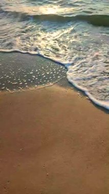 Gün batımında deniz kıyısındaki kumlu sahilde dalgalar dalgalanır. Beyaz köpüklü mavi deniz dalgaları kumsala geliyor. Deniz kıyısı kıyı şeridi. Doğal arka plan, doğa. Dikey