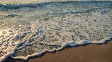 Gün batımında deniz kıyısındaki kumlu sahilde dalgalar dalgalanır. Beyaz köpüklü mavi deniz dalgaları kumsala geliyor. Deniz kıyısı kıyı şeridi. Doğal zemin, doğa zemin