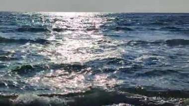 Mavi deniz ve güneş güneşli yaz gününde deniz dalgalarının yüzeyinde parlar. Güneşli bir günde kumlu sahil sahillerinde dalgalar halinde akan mavi deniz. Beyaz köpük, deniz kuzuları. Doğal arkaplan.