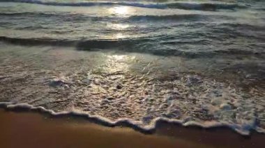 Gün batımında deniz kıyısındaki kumlu sahilde dalgalar dalgalanır. Beyaz köpüklü mavi deniz dalgaları kumsala geliyor. Deniz kıyısı kıyı şeridi. Doğal zemin, doğa zemin