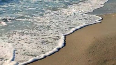 Mavi deniz, kumlu sahil ve güneşli yaz gününde deniz dalgalarının yüzeyinde parlayan güneş. Güneşli bir günde kumlu sahil sahillerinde dalgalar halinde akan mavi deniz. Beyaz köpük, deniz kuzuları. Doğal arkaplan.