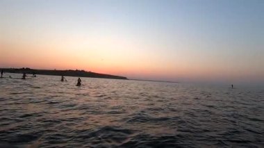 Şafak vakti SUP sörf tahtasında yüzen bir adam. Beyaz SUP tahtasının üzerinde yüzen ve günbatımında kıyıya yakın dalgalarla kürek çeken insanlar. Aktif yaşam tarzı eğlence sporu POV