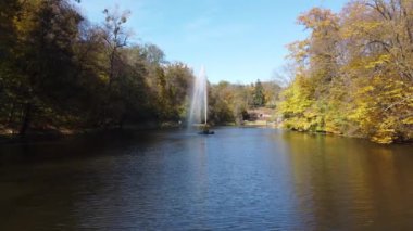 Güneşli bir sonbahar gününde gölün ortasında dekoratif fıskiyeler, ağaçlar ve büyük taşlar olan bir park. Bir sürü turist. Düşen yapraklar ve mavi gökyüzü yansıması ile suyun üzerinde uçmak. Güzel manzara.