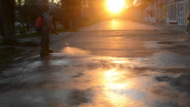 身着紧身衣的男子在日落的背景下在城市的街道上喷洒消毒液 清洁工人清扫人行道 卫生措施 Coronavirus大流行病 — 图库视频影像