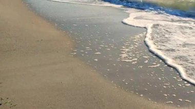 Mavi deniz, kumlu sahil ve güneşli yaz gününde deniz dalgalarının yüzeyinde parlayan güneş. Güneşli bir günde kumlu sahil sahillerinde dalgalar halinde akan mavi deniz. Beyaz köpük, deniz kuzuları. Doğal arkaplan.