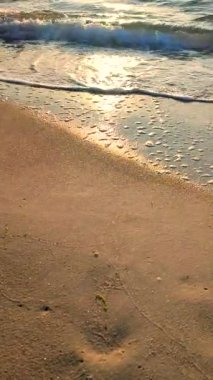 Gün batımında deniz kıyısındaki kumlu sahilde dalgalar dalgalanır. Beyaz köpüklü mavi deniz dalgaları kumsala geliyor. Deniz kıyısı kıyı şeridi. Doğal arka plan, doğa. Dikey