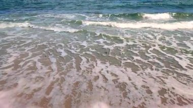 Mavi deniz, dalgalar, beyaz köpük, kumlu sahil, güneşli yaz gününde kıyılar. Beyaz köpüklü mavi deniz dalgaları kumlu deniz kıyısında güneşli bir güne çarpıyor. Deniz manzarası deniz manzarası manzarası manzarası. Yavaş çekim