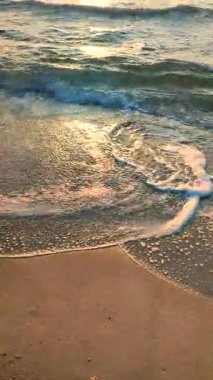 Gün batımında deniz kıyısındaki kumlu sahilde dalgalar dalgalanır. Beyaz köpüklü mavi deniz dalgaları kumsala geliyor. Deniz kıyısı kıyı şeridi. Doğal arka plan, doğa. Dikey