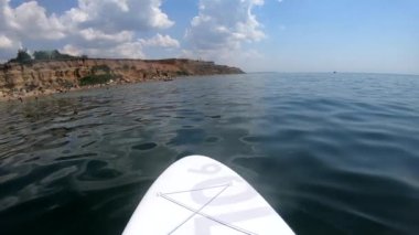 Güneşli bir yaz gününde kumsalın kıyısında mavi denizde beyaz bir SUP tahtasında yüzen biri. Okyanus gölünde su yüzeyinde yiyecek. Bakış açısı. Geniş açı. Aktif yaşam tarzı istirahat rahatı. Yavaş çekim