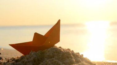 Paper orange boat on the sand near the sea waves with sunny path on sandy beach of sea shore during sunset dawn with shining sun close-up. Swimming dream sailing traveling vacation tourism concept