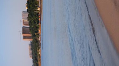 Deniz kıyısı yazın erken saatlerinde. Mavi deniz, deniz kenarındaki kumlu sahil yüksek binalar ve güneşli bir yaz sabahı yeşil ağaçlar.