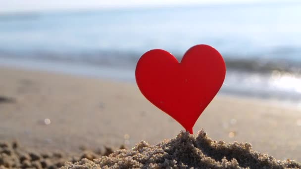 晴れた夏の日の終わりに海の波の背景に海岸の砂浜の砂の赤い心の形をしたプラスチック棒 コンセプト 恋愛関係 ロマンス アムール セント バレンタインデー — ストック動画