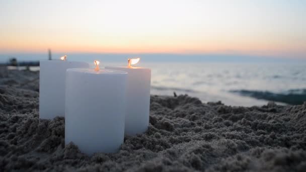 3つの大きなパラフィン白いろうそくは日没と夜明けのクローズアップで海岸海岸の海の波の近くの砂浜で燃えています コンセプト ロマンチックな気分 ロマンスデート お祝い パーティー — ストック動画