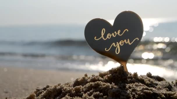 太陽の晴れた夏の日の終わりに海の波の海岸の砂浜であなたを愛する言葉の心のプラスチック棒の形 コンセプト 恋愛関係 ロマンス アムールのお祝い セントバレンタインデー — ストック動画