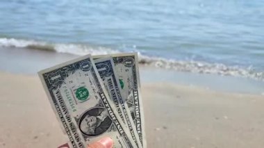 Elinde, güneşli bir yaz gününde kumlu sahildeki deniz dalgalarının arka planında bir dolar değerindeki Amerikan dolarının üç banknotunu tutan kişi. Para finansmanı kavramı