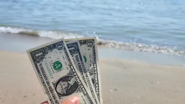 晴れた夏の日に海岸の砂浜で海の波の背景に1ドルの顔の価値を持つ米ドル紙幣の3つの宗派を手に持っている人 コンセプトマネーファイナンス — ストック動画