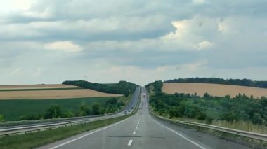 Yol boyunca giden bir arabanın ön camından geçen arabaların bulunduğu asfalt çok şeritli bir yol manzarası. Otoyol manzarası, sarı buğday tarlaları, ağaçlar ve bulutlu gökyüzü. Manzara Manzarası