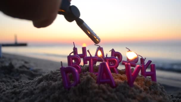 男は夜明けの日の海岸の砂浜のピンク色のハッピーバースデーの文字の形でガスバーナーでろうそくを点灯します 海岸でろうそくを燃やした コンセプトのお祝いパーティー — ストック動画