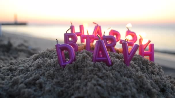 日没の夜明けのクローズアップ中に海岸の砂浜に燃えているピンク色のハッピーバースデーの文字の形のろうそく 多くのろうそくが海の近くで砂の上に燃えている コンセプトのお祝いパーティー — ストック動画