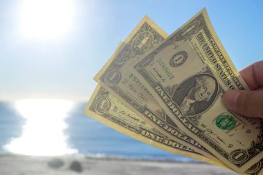 Elinde, güneşli bir yaz gününde kumlu sahildeki deniz dalgalarının arka planında bir dolar değerindeki Amerikan dolarının üç banknotunu tutan kişi. Para finansmanı kavramı