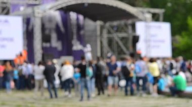 Arka plan bulanık. Sahnede performans. Sahnenin yanında duran bir sürü insan. İnsanlar doğada sahneye çıkıyorlar. Sahnedeki konser. Doğadaki kapalı mekan sahnesinin yanında bir sürü insan duruyor.