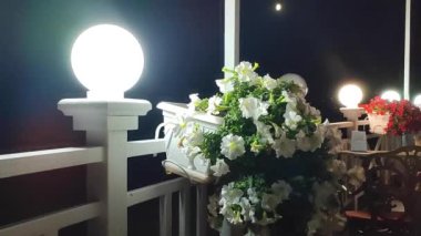 Karanlık bir gecede parlayan fener ve beyaz çiçek açan petunya kahve masasının yanındaki çitte sallanan rüzgarda sallanan beyaz tencerede sallanıyor. Gece kafede romantik bir atmosfer. İç dekorasyon ayrıntıları dışarıda.
